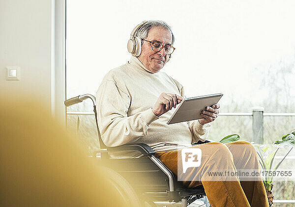 Senior disabled man wearing wireless headphones using tablet PC on wheelchair