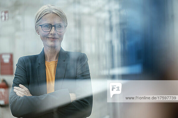 Confident businesswoman with arms crossed seen through glass of office
