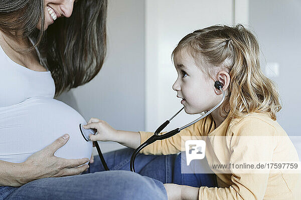 Curious girl listening to pregnant mother's belly through stethoscope