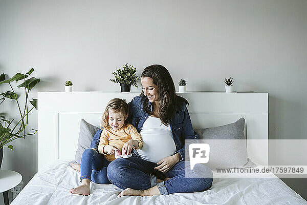 Happy pregnant woman looking at daughter holding baby booties on bed