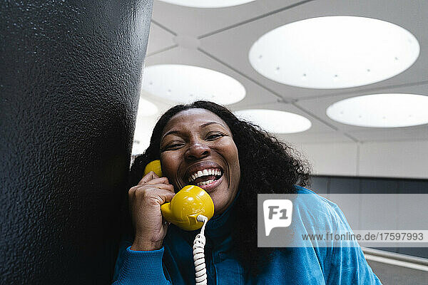 Happy woman listening through old-fashioned yellow telephone receiver leaning on column