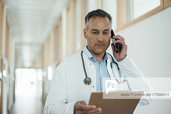 Mature doctor examining medical record talking on smart phone in hospital