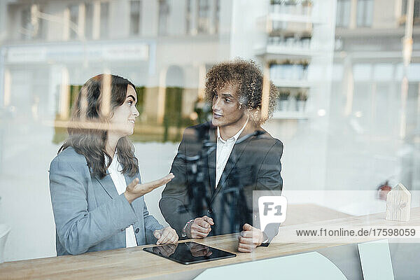 Young businesswoman discussing with businessman at desk in coworking office seen through glass