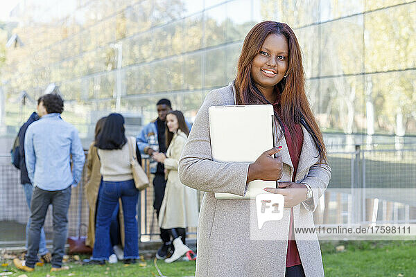 Redhead student holding laptop with friends in background at college campus