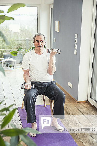 Senior man with dumbbells sitting on chair working out at home