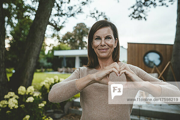 Smiling woman gesturing heart shape at backyard
