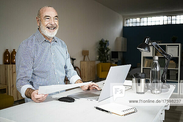 Cheerful businessman with document and laptop sitting at desk