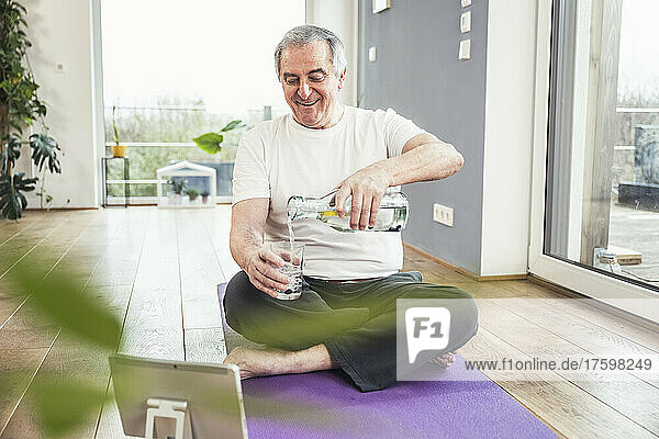 Happy senior man sitting on exercise mat pouring water in drinking glass at home