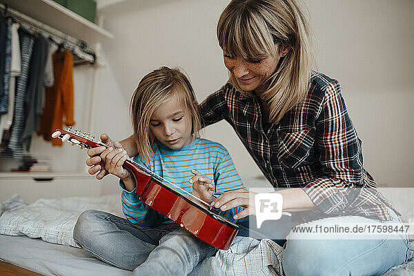 Mother teaching son to play guitar in bedroom at home