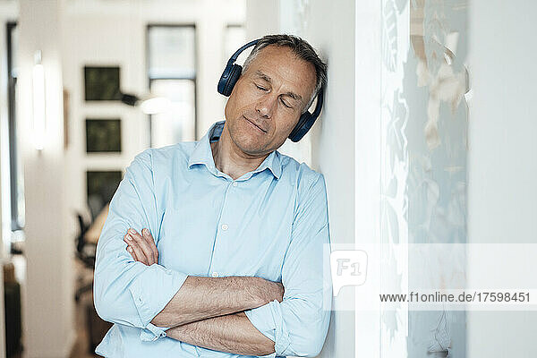 Businessman with eyes closed listening music and leaning on wall in office