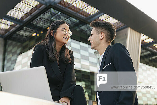 Smiling businesswoman with laptop looking at colleague in front of office building