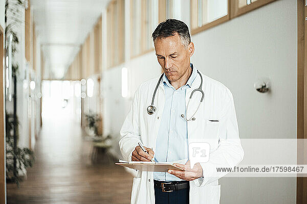 Mature doctor writing medical record at corridor in hospital