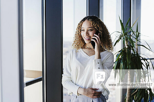 Young businesswoman with curly hair talking on smart phone near window in office