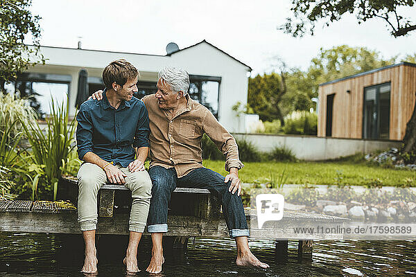 Älterer Mann unterhält sich mit seinem Sohn  der auf einem Steg am See im Hinterhof sitzt