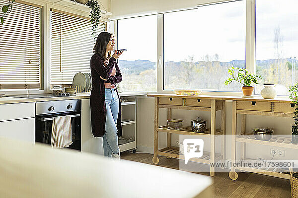 Young woman talking on speaker phone leaning on kitchen counter at home