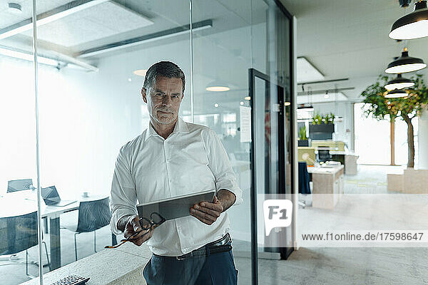 Confident businessman with tablet PC leaning on glass wall at office