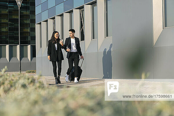 Businessman with trolley bag walking with colleague on footpath by office building