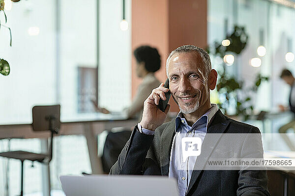 Lächelnder Geschäftsmann  der im Büro auf dem Smartphone spricht