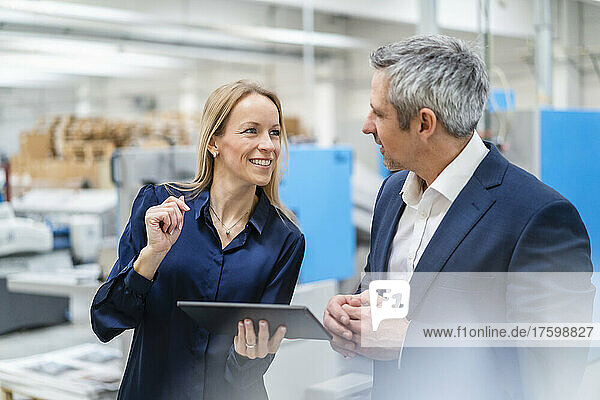 Happy blond businesswoman holding tablet PC discussing with colleague in factory