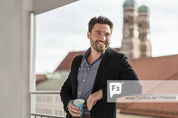 Smiling businessman with coffee cup leaning on railing at office balcony