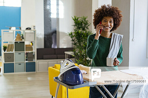 Happy Afro woman talking on phone standing at ironing board