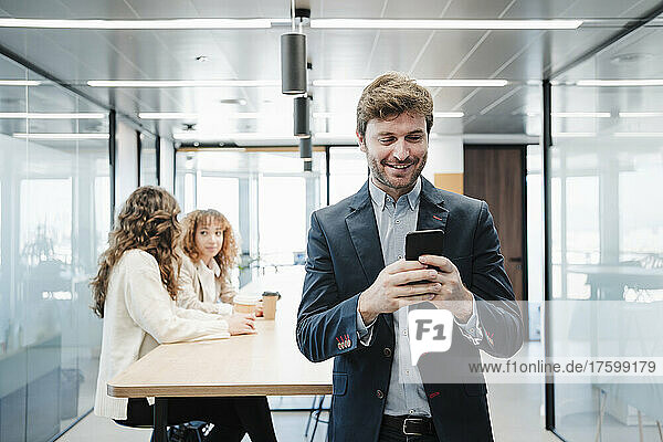 Lächelnder Geschäftsmann  der im Büro Textnachrichten über sein Smartphone sendet