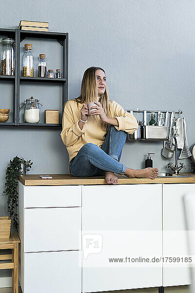 Thoughtful woman with coffee cup sitting on kitchen counter