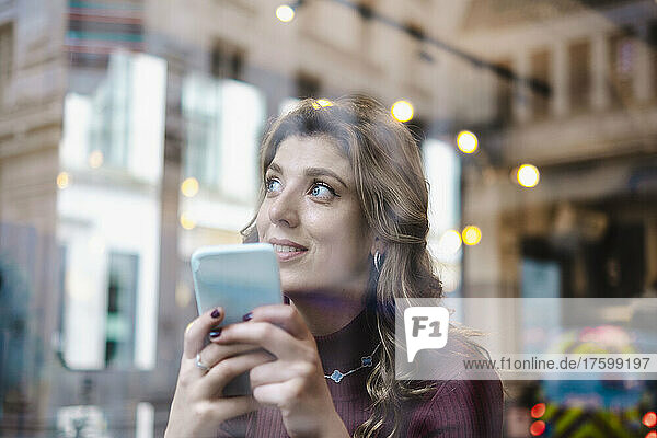 Thoughtful woman with smart phone seen through window of cafe