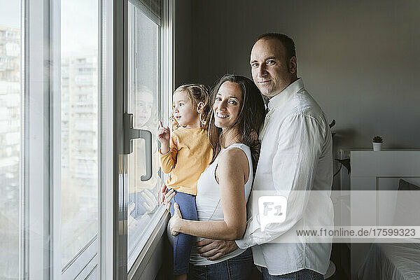 Lächelnde Eltern stehen mit ihrer Tochter und schauen zu Hause durch das Fenster