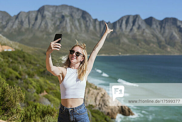Lächelnde junge Frau mit erhobener Hand macht an einem sonnigen Tag ein Selfie mit dem Mobiltelefon