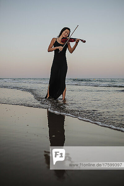 Frau spielt Geige und steht bei Sonnenuntergang im Meer