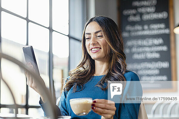 Smiling woman with coffee cup on video call over mobile phone in cafe