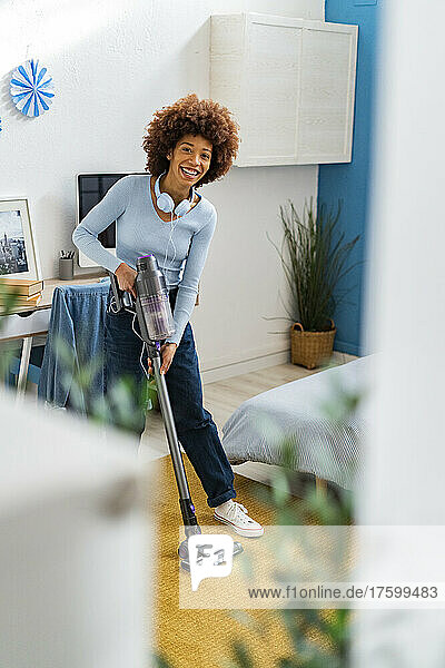 Smiling young Afro woman cleaning carpet with vacuum cleaner in bedroom at home