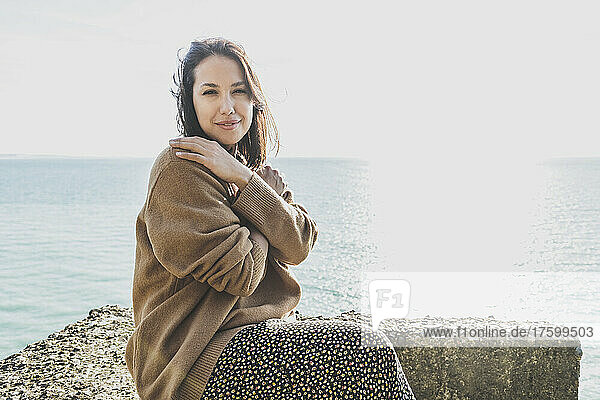 Beautiful young woman hugging self sitting on rock at beach