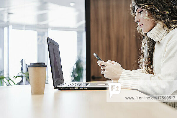 Businesswoman using smart phone over laptop at desk in office
