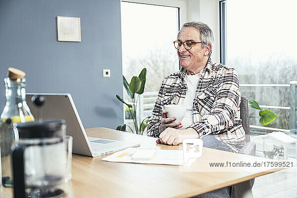 Smiling senior man holding coffee cup watching videos on laptop at home