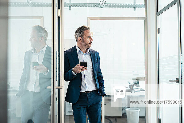 Thoughtful businessman leaning with hand in pocket holding smart phone at office