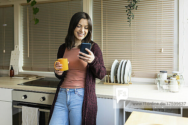 Smiling woman using smart phone holding mug leaning on kitchen counter at home