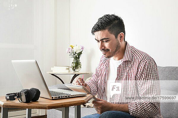 Smiling man writing in notepad by laptop in living room