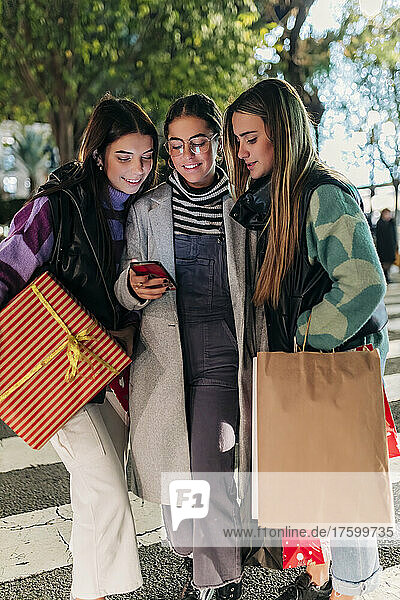 Young friends with shopping bags sharing smart phone on street