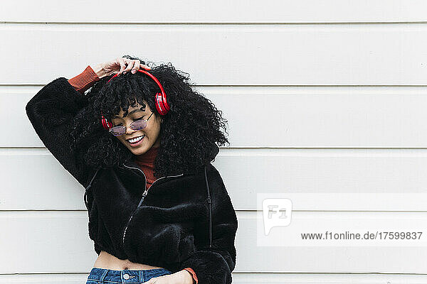 Happy young fashionable woman listening music through headphones in front of white wall