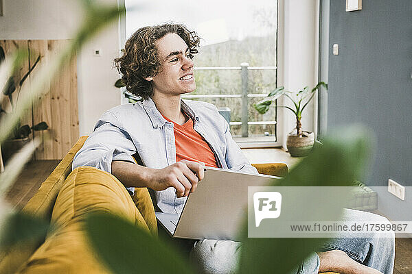 Thoughtful smiling man with laptop sitting on sofa in living room at home
