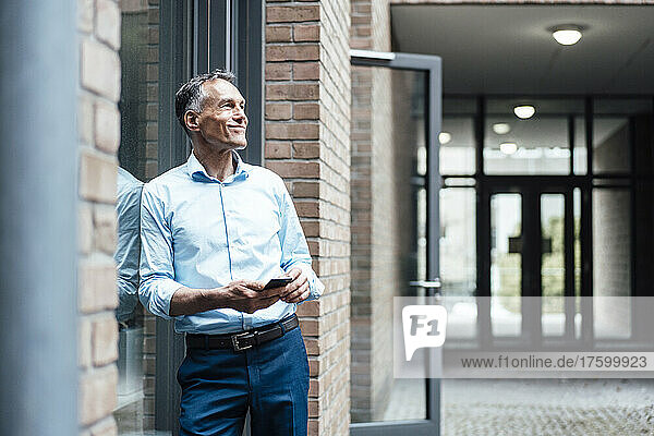Confident mature businessman leaning on window outside office building