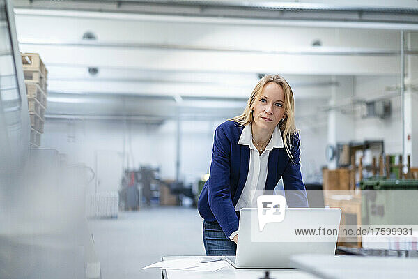 Thoughtful blond businesswoman with laptop leaning on desk in factory