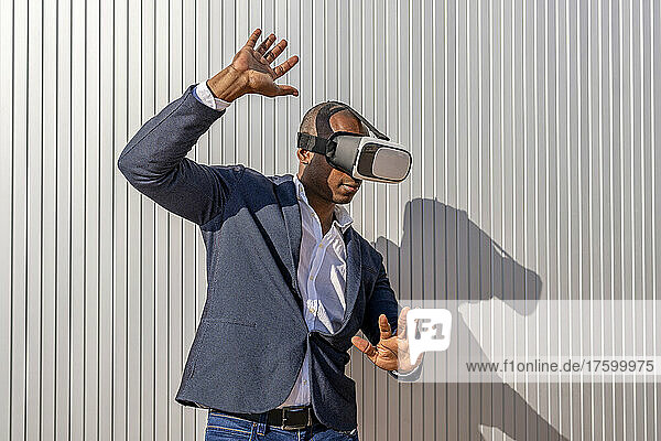 Businessman wearing virtual reality simulator gesturing in front of wall on sunny day