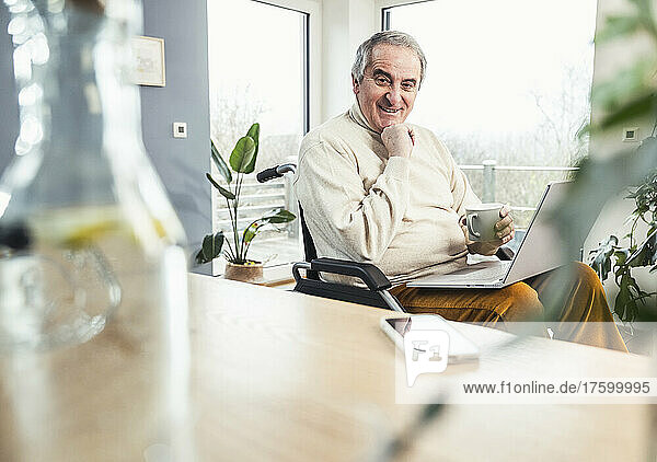 Smiling disabled man holding coffee cup sitting with hand on chin at home