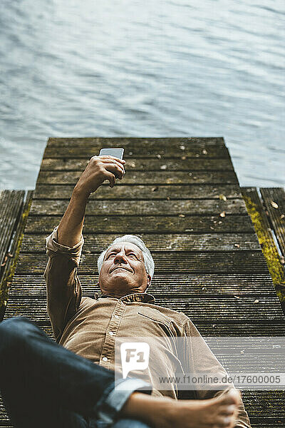 Senior man taking selfie through smart phone lying on jetty by lake