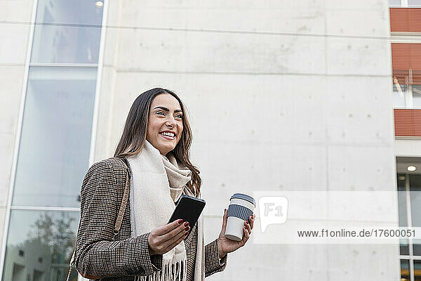 Lächelnde junge Frau mit Smartphone und wiederverwendbarer Kaffeetasse vor dem Gebäude