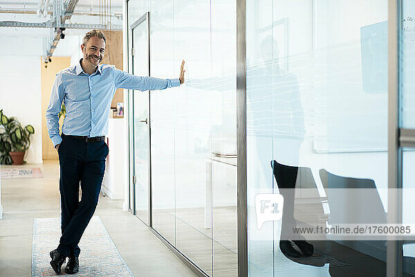 Smiling businessman leaning on glass wall in corridor at workplace