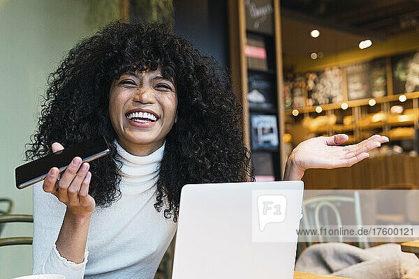 Cheerful young woman sitting with smart phone and laptop gesturing at cafe